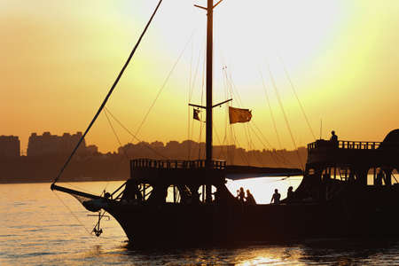 Silhouette of a beautiful ship with balks, lowered sails, waving flags and a raised anchor with many people on board on the background of bright warm sunset in the sea near the shoreline in evening.の写真素材