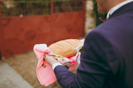 Wedding traditions Bride buying The groom in a dark purple suit and white shirt with a bow-tie and boutonniere holds a bread loaf on a napkin outdoors near the brown gate.の写真素材