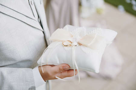 Close up Two beautiful stylish gold wedding rings of the bride and groom are on a refined white pillow with lace and bows in the hands of a man in a gray suit. Wedding accessories, jewelry.の写真素材