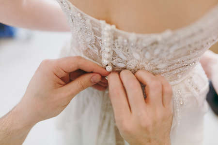 Close up Morning preparation newlyweds for the wedding. Men's hands button up elegant lace delicate ivory wedding dress with beads at the waist for bride. Wedding wear, accessories.の写真素材