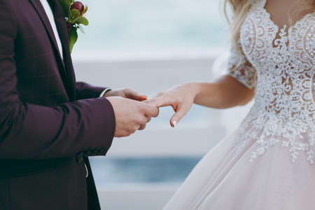 Beautiful wedding celebration. A stylish bridegroom in a dark purple suit holds the hand and puts the ring on his bride in a white elegant dress with rhinestones outdoors. Wedding wear, accessoriesの写真素材