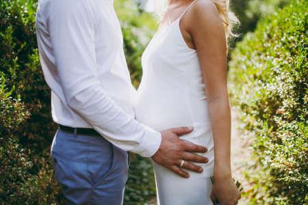 Beautiful wedding celebration. The groom in gray trousers and a white shirt hugs his pregnant blonde bride in an elegant simple white dress in the middle of the garden on a sunny day.の写真素材