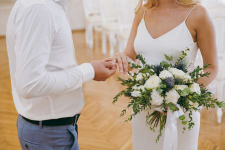 Beautiful wedding celebration. The groom in gray trousers and a white shirt puts engagement ring his pregnant blonde bride in simple white dress with a bouquet of light flowers and green twigs.の写真素材