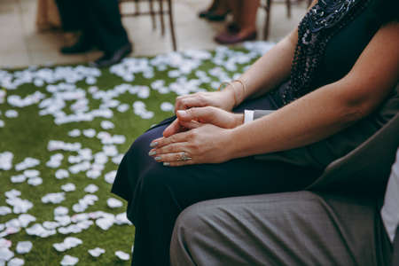 Beautiful wedding celebration. Elderly couple in formal clothes sitting on chairs and holding hands at wedding ceremony near a path covered with petals of flowers in the open airの写真素材