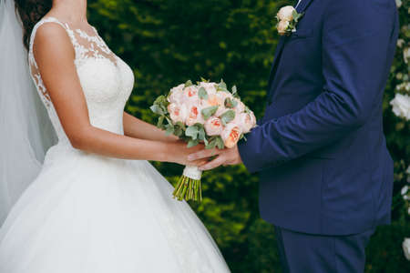 Beautiful wedding celebration. The groom in a blue suit with a boutonniere and the bride in an elegant lace dress with veil and a bouquet of peonies and green leaves holding hands in the green garden.の写真素材