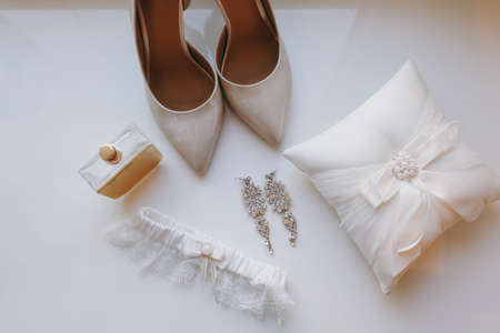Close up Elegant light beige wedding varnish shoes with sharp noses, pillow for rings, earrings, perfume and garter for the bride on a white gloss windowsill background. Wedding wear, accessories.の写真素材