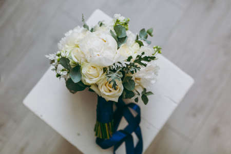 Close up A beautiful tender bridal bouquet in pastel colors of roses, peonies and green twigs tied with a long silk blue ribbon on background a wooden chair. Wedding accessories for bride.の写真素材