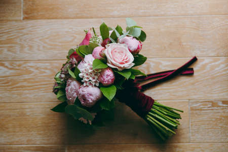 Close up A beautiful tender bridal bouquet in rose colors of roses, peonies and green twigs tied with a long velvet claret ribbon on a background of a wooden floor. Wedding accessories for bride.の写真素材