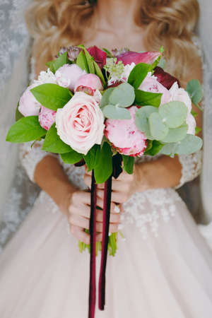 Close up A beautiful tender bridal bouquet in rose colors of roses, peonies and green twigs tied with a long velvet claret ribbon in the hands of the bride. Wedding accessories for bride.の写真素材