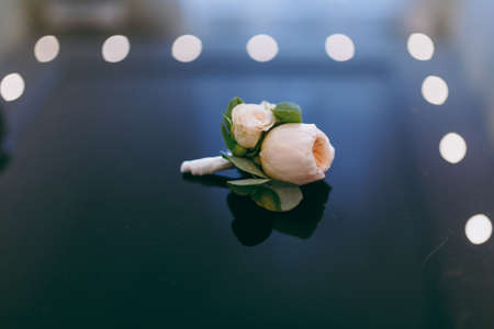 Close up A beautiful tender wedding boutonniere for the groom of light roses and green leaves with white silk ribbon on a dark glossy background with glare from light bulbs. Wedding accessories.の写真素材