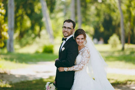 Beautiful wedding photosession. The groom in a black suit and his bride in a white lace dress with a long plume, veil and bouquet smile and embrace in a large green garden on weathery sunny day.の写真素材