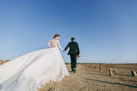 Beautiful wedding photosession. Handsome unshaved groom in a black trousers with bouquet and young cute bride in white lace pattern dress with exquisite hairstyle on walk along the coastline.の写真素材