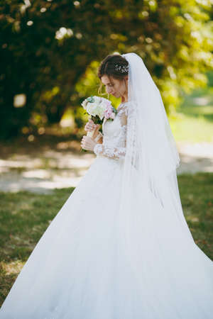 Beautiful wedding photosession. The bride in a white lace dress with a long plume, hairpin in hair, veil and bouquet of white and pink flowers in a large green garden on weathery sunny day.の写真素材