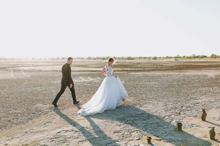 Beautiful wedding photosession. Handsome unshaved groom in a black trousers with bouquet and young cute bride in white lace pattern dress with exquisite hairstyle on walk along the coastline sunset.の写真素材