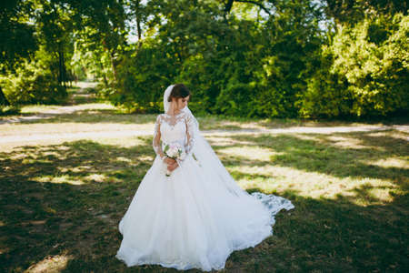 Beautiful wedding photosession. The bride in a white lace dress with a long plume, hairpin in hair, veil and bouquet of white and pink flowers in a large green garden on weathery sunny day.の写真素材