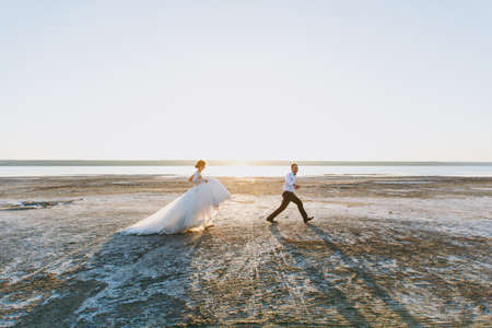Beautiful wedding photosession. Handsome unshaved groom in a black trousers and young cute bride in white lace pattern dress with exquisite hairstyle on walk along the coastline near the sea sunset.の写真素材