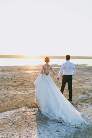 Beautiful wedding photosession. Handsome unshaved groom in a black trousers and young cute bride in white lace pattern dress with exquisite hairstyle on walk along the coastline near the sea sunset.の写真素材
