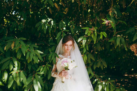 Beautiful wedding photosession. Young tender cute smiling bride in elegant lace dress with long sleeves, veil and bouquet of white and pink flowers amidst the branches of green bush in garden.の写真素材