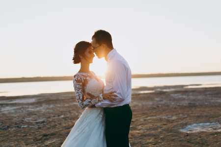 Beautiful wedding photosession. Handsome unshaved groom in a black trousers and young cute bride in white lace pattern dress with exquisite hairstyle on walk along the coastline near the sea sunset.の写真素材