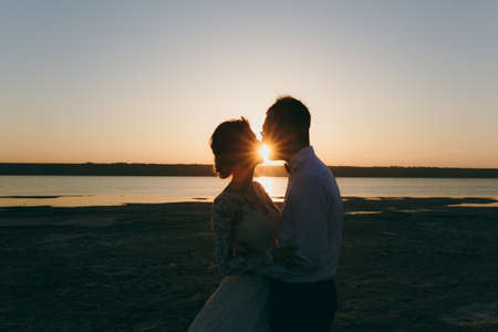 Beautiful wedding photosession. Handsome unshaved groom in a black trousers and young cute bride in white lace pattern dress with exquisite hairstyle on walk along the coastline near the sea sunset.の写真素材