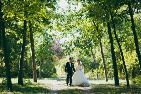 Beautiful wedding photosession. The groom in a black suit with bouquet and his bride in a white lace dress with a long plume and veil smile and walk along the path in large green garden on sunny day.の写真素材