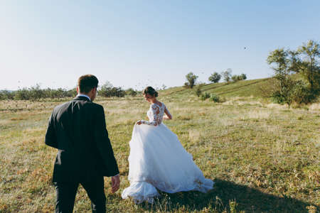 Beautiful wedding photosession. Handsome groom in a black suit and young bride in white lace dress with exquisite hairstyle walk around the big green field against the sky and flying birds.の写真素材