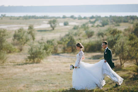 Beautiful wedding photosession. Handsome groom in a black suit and young bride in white lace dress with exquisite hairstyle on walk around the big green field against the trees and bushes background.の写真素材