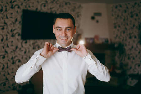 Morning preparation of the newlyweds for the wedding ceremony. The groom in a white shirt with bracelet on his hand straightens a patterned bow tie on his collar and smiles. Wedding wear, accessories.の写真素材