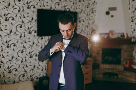 Morning preparation of the newlyweds for the wedding ceremony. The groom in white shirt, blue trousers, jacket and bow tie puts on cufflinks on sleeves in room with patterned wallpaper. Wedding wear.の写真素材