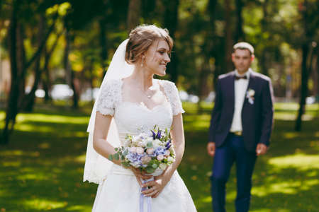 Beautiful wedding photosession. Handsome groom in blue formal suit and his elegant bride in dress and veil with bouquet with beautiful hairdress embrace on a walk in the big green park on sunny day.の写真素材