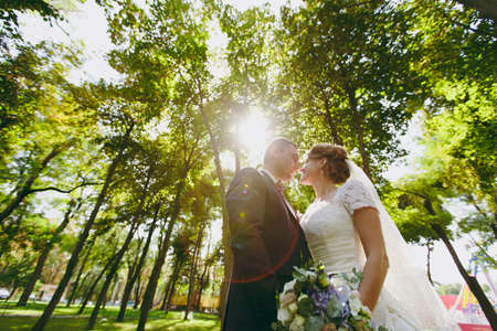 Beautiful wedding photosession. Handsome groom in blue formal suit with bouquet and his elegant bride in white dress and veil with beautiful hairdress on a walk in the big green park on a sunny day.の写真素材