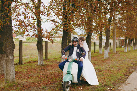Beautiful wedding photosession. Handsome groom in blue formal suit and helmet with boutonniere and his elegant bride in white dress and veil amusing themselves on a green scooter near autumn trees.の写真素材