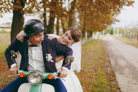 Beautiful wedding photosession. Handsome groom in blue formal suit and helmet with boutonniere and his elegant bride in white dress and veil amusing themselves on a green scooter near autumn trees.の写真素材