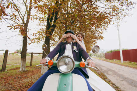 Beautiful wedding photosession. Handsome groom in blue formal suit and helmet with boutonniere and his elegant bride in white dress and veil amusing themselves on a green scooter near autumn trees.の写真素材