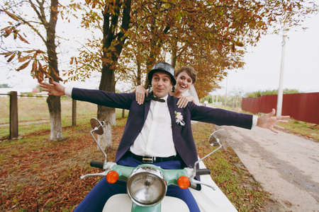 Beautiful wedding photosession. Handsome groom in blue formal suit and helmet with boutonniere and his elegant bride in white dress and veil amusing themselves on a green scooter near autumn trees.の写真素材