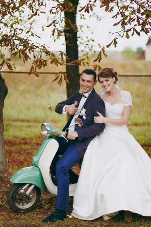 Beautiful wedding photosession. Handsome groom in blue formal suit and bow tie with boutonniere and his elegant bride in white dress and veil sit on a green vintage scooter near autumn trees.の写真素材