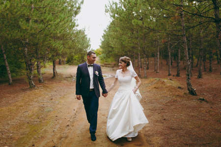 Beautiful wedding photosession. Handsome groom in blue formal suit and bow tie with boutonniere and his elegant bride in white dress and veil with a beautiful hairdress on a walk in coniferous forest.の写真素材
