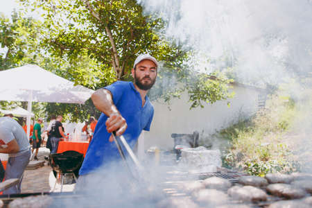 Odessa, Ukraine, 08-26-2017:A bearded tanned man in a blue T-shirt and cap prepares and turns over hot meat puffing steaks on grill with forceps at a barbeque party outdoors. Concept of lifestyle.のeditorial素材