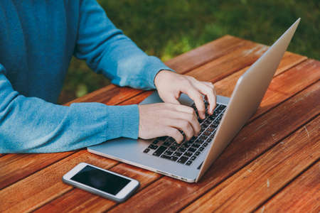 Close up cropped portrait of successful smart man businessman or student sitting at table with mobile phone in city park using laptop, working outdoors. Mobile Office concept. Hands on the keyboardの写真素材