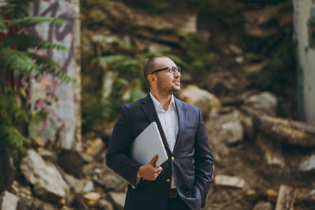 Young successful smart businessman in white shirt, classic suit, glasses. Man standing with laptop...