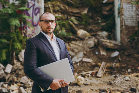 Young successful smart businessman in white shirt, classic suit, glasses. Man standing with laptop pc computer phone near ruins, debris, stone building outdoors. Mobile Office, business, work conceptの写真素材