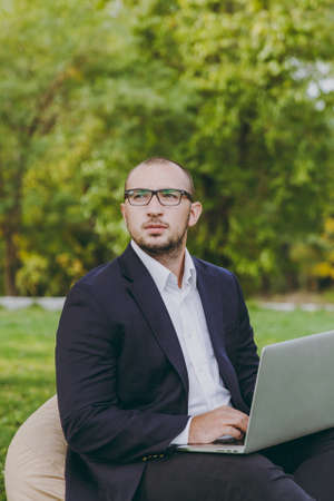Young successful businessman in white shirt, classic suit, glasses. Man sit on soft pouf, working on laptop pc computer in city park on green lawn outdoors on nature. Mobile Office, business conceptの写真素材