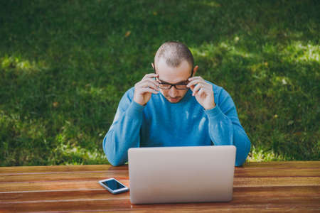 Young successful smart man businessman or student in casual blue shirt glasses sitting at table with mobile phone in city park using laptop working outdoors on green nature. Mobile Office conceptの写真素材