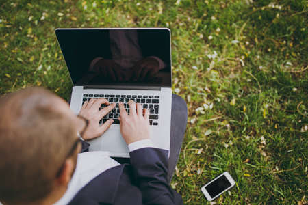 Close up cropped businessman in classic suit. Man sit on soft pouf, working on laptop pc computer in city park on green lawn outdoors on nature. Mobile Office, business concept. Top view. Copy spaceの写真素材