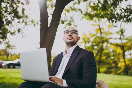 Young successful businessman in white shirt, classic suit, glasses. Man sit on soft pouf, working on laptop pc computer in city park on green lawn outdoors on nature. Mobile Office, business conceptの写真素材