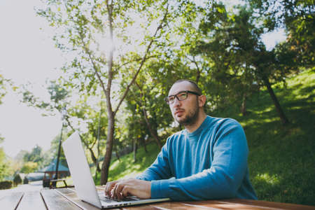 Young successful smart man businessman or student in casual blue shirt glasses sitting at table with mobile phone in city park using laptop working outdoors on green nature. Mobile Office conceptの写真素材