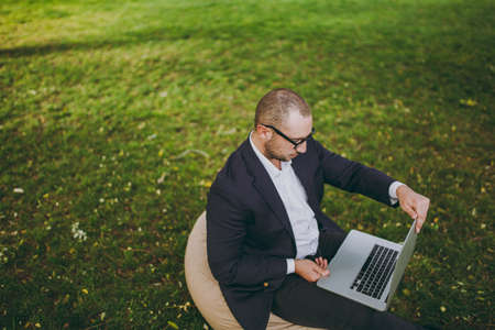 Young successful businessman in white shirt, classic suit, glasses. Man sit on soft pouf, working on laptop pc computer in city park on green lawn outdoors on nature. Mobile Office concept. Top viewの写真素材