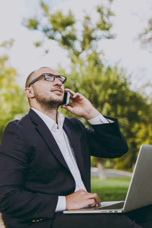 Young businessman in white shirt, classic suit, glasses. Man sit on soft pouf, work on laptop pc computer, talk on mobile phone in city park on green lawn outdoors. Mobile Office, business conceptの写真素材