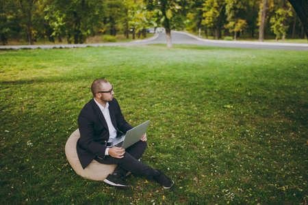 Young successful businessman in white shirt, classic suit, glasses. Man sit on soft pouf, working on...