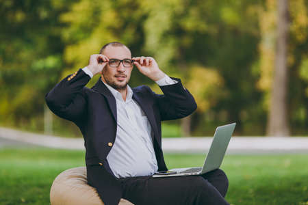 Successful businessman in white shirt, classic suit, correct glasses. Man sit on soft pouf, work on laptop pc computer in city park on green lawn outdoors on nature. Mobile Office, business conceptの写真素材
